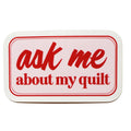 A rectangular sign with rounded corners featuring the text 'ask me about my quilt' in red script and serif lettering on a white background with a red border.