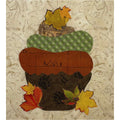 A square art print or textile panel featuring a stylized acorn made from layered autumn-themed fabrics and paper. The acorn has a brown earth-like base with dark brown paw-print texture, a medium brown section with mountain line art, a green diamond-patterned upper section, and yellow/orange leaf accents.