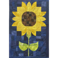 A square quilted wall hanging or blanket featuring a large yellow sunflower with a brown patterned center, green stem, and blue patchwork background.