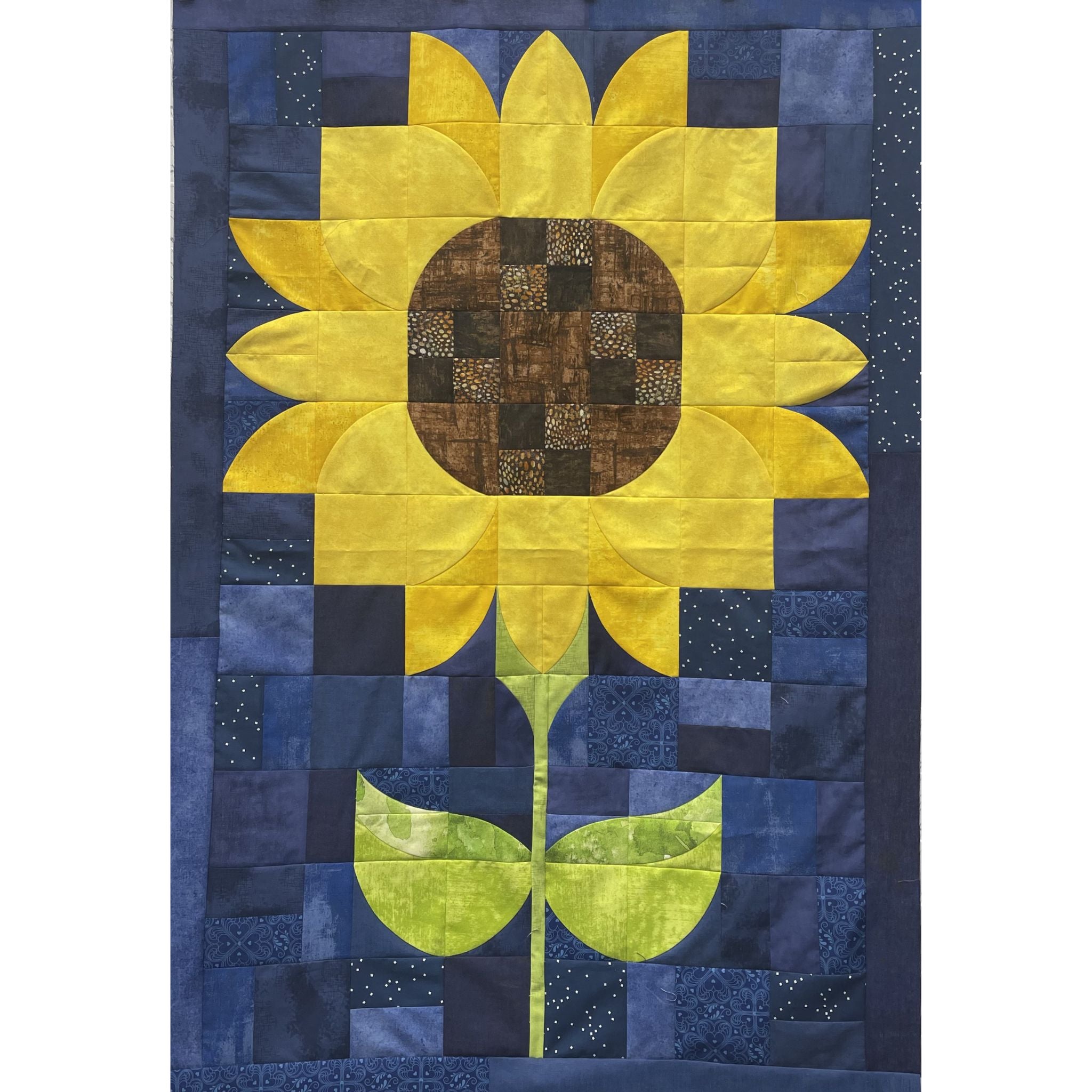 A square quilted wall hanging or blanket featuring a large yellow sunflower with a brown patterned center, green stem, and blue patchwork background.