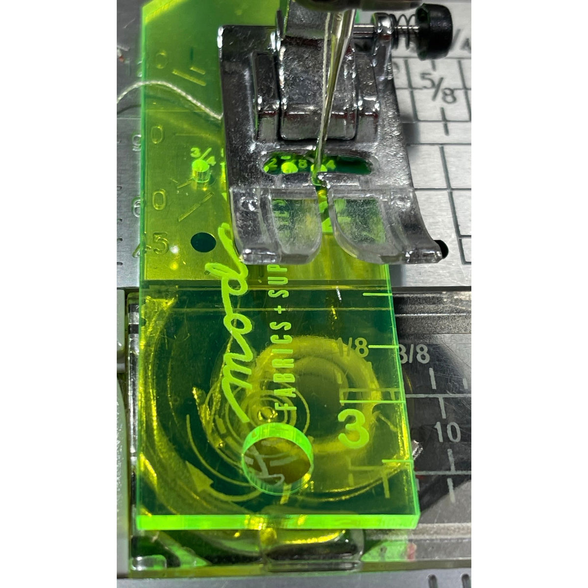 A close-up of a sewing machine foot and presser bar assembly with a bright neon green transparent guide block. The guide has white printed markings and the word 'model' in white script.